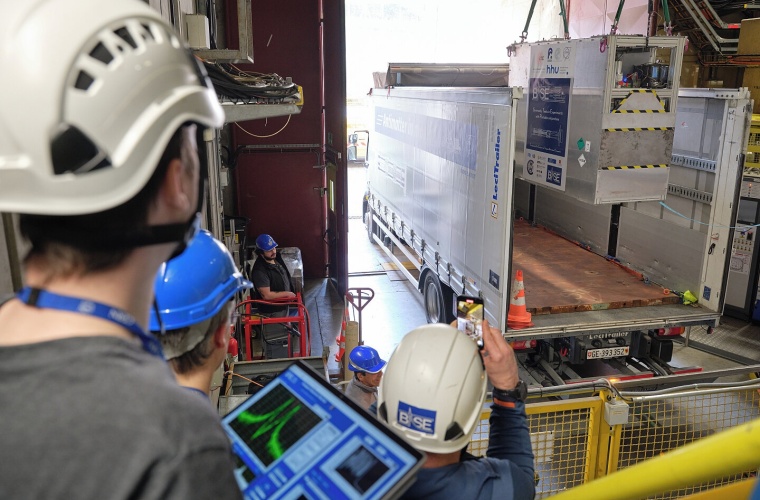 Die Falle BASE-STEP wird vom Kran in den Transport-Lkw gehoben; im Vordergrund HHU-Doktorand Marcel Leonhardt mit dem mobilen Kontrollterminal für die Falle. (Foto: CERN)