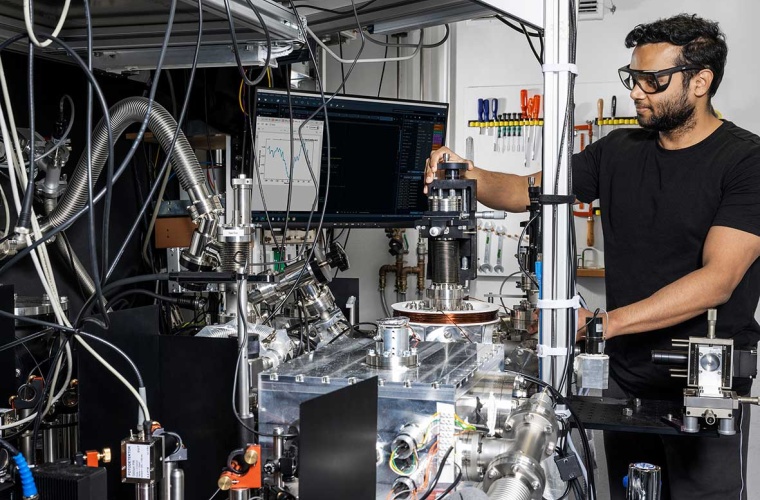 Vishal Lal mit dem Kernuhr-Experiment der PTB. Auf dem optischen Tisch befindet sich der Aufbau für die Erzeugung des vakuumultravioletten Laserlichts und die Vakuumkammer mit dem Th-dotierten Kristall