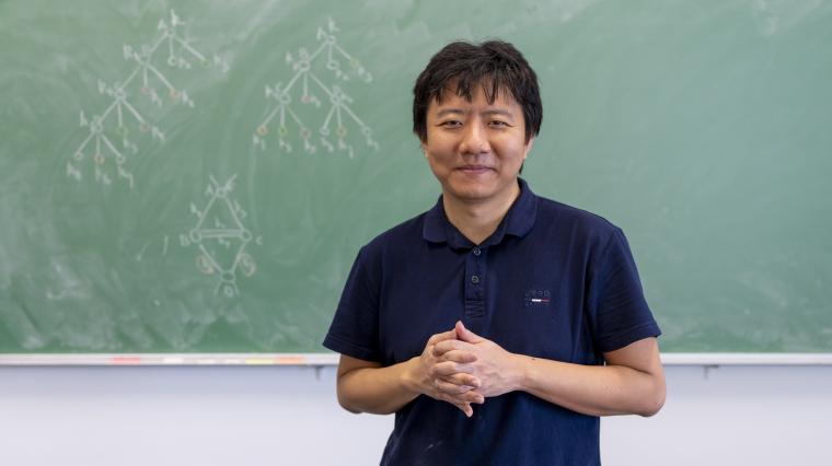 Yu Deng steht vor einer grünen Tafel, auf der Baumdiagramme zu sehen sind.